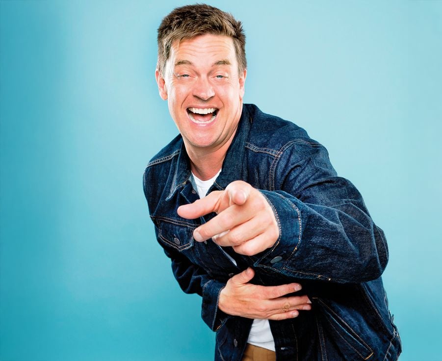 A man wearing a denim jacket is laughing and pointing toward the camera, holding his stomach with his other hand, in front of a plain blue background.