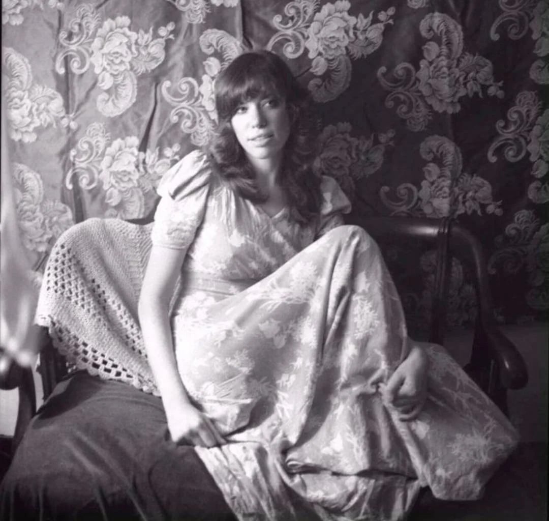 A woman in a long, patterned dress sits on a vintage couch with a crocheted blanket, surrounded by ornate floral wallpaper. She gazes at the camera with a relaxed expression in this black-and-white photo.
