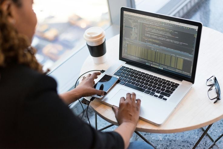 A person sits at a round table working on a laptop with code on the screen. A smartphone, charging cables, and a takeaway coffee cup are also on the table near a large window.