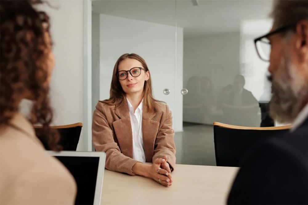 A woman in glasses and a brown blazer sits at a table, facing two people during an interview or meeting in a modern office setting.