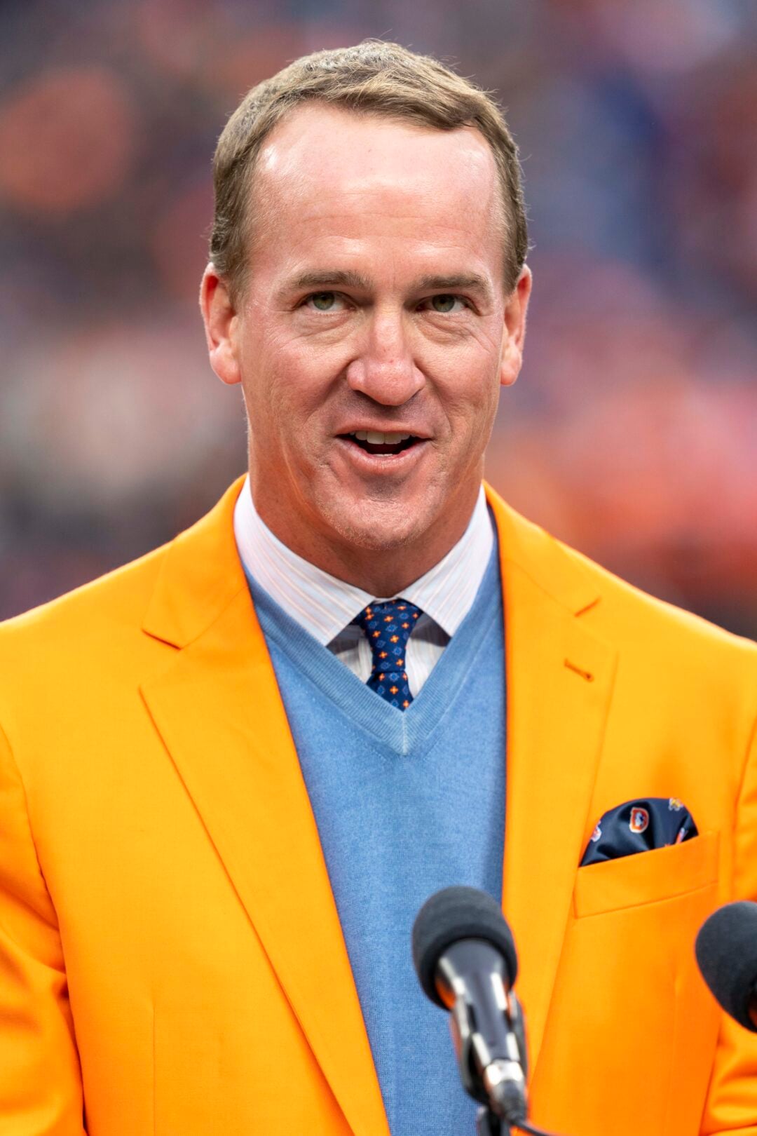A man wearing a bright orange blazer, light blue sweater, white shirt, and patterned tie stands in front of microphones, speaking in an outdoor setting with a blurred crowd in the background.
