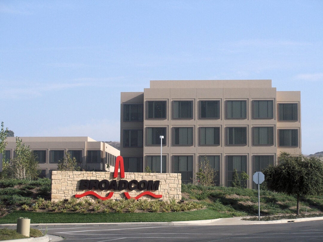 A modern office building with large windows is seen behind a landscaped area featuring a stone sign that reads "Broadcom" in red and black letters. Trees and shrubs are planted around the sign.