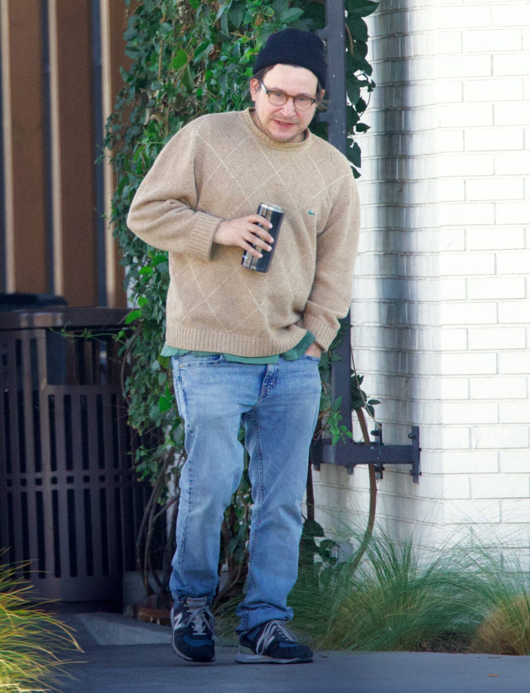 A person wearing a beige sweater, blue jeans, black sneakers, black beanie, and glasses is standing outside near a white brick wall, holding a beverage can and looking at the camera.