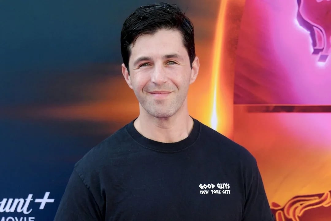 A man with short dark hair wearing a black "Good Guys New York City" t-shirt smiles at the camera, standing in front of a colorful, illuminated background.