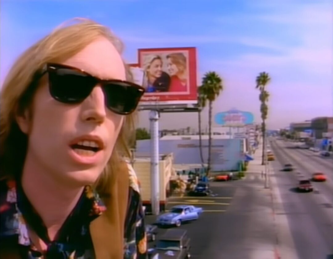 A man wearing sunglasses and a patterned shirt stands on a city street with palm trees, cars, and billboards in the background on a sunny day.
