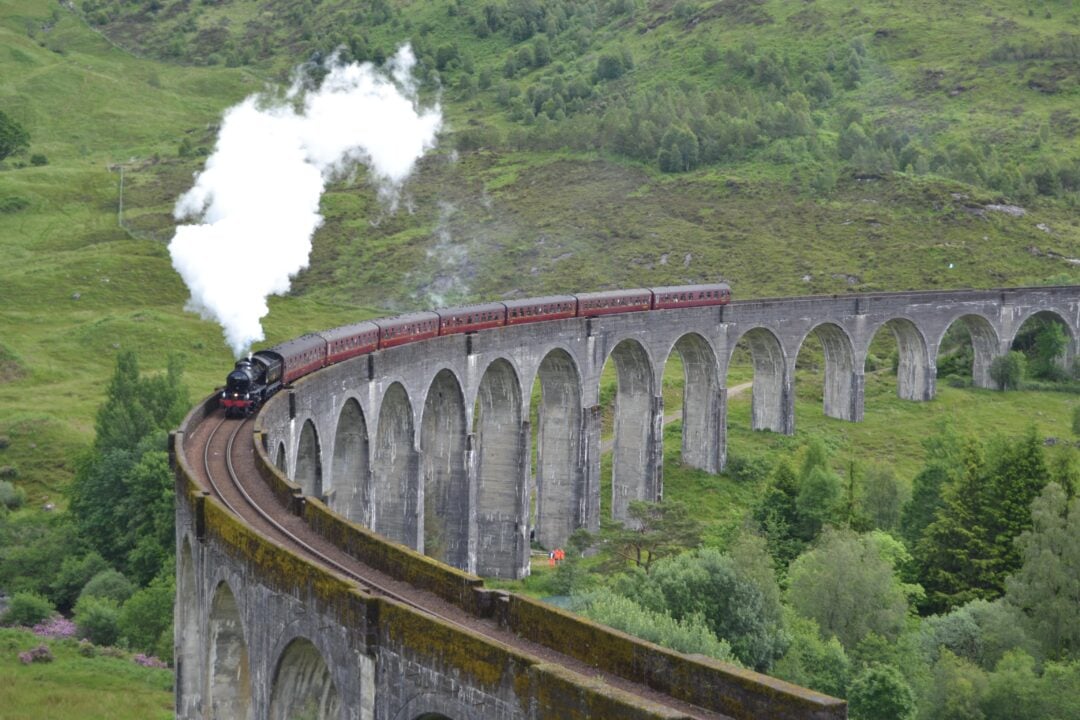 A steam train with red carriages travels across a large, curved stone viaduct surrounded by lush green hills, with white smoke billowing from its engine.