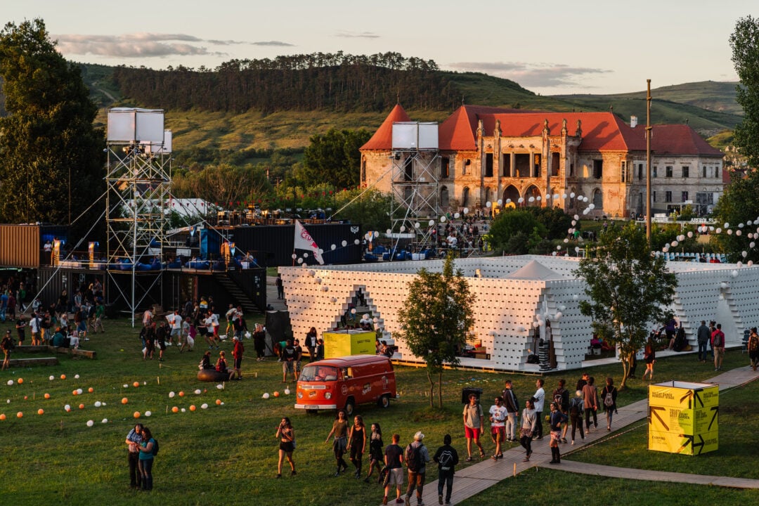 A lively outdoor festival takes place near a historic castle, with people walking, tents and structures set up, colorful lights, and a red van parked on the grass. Green hills and trees surround the area.