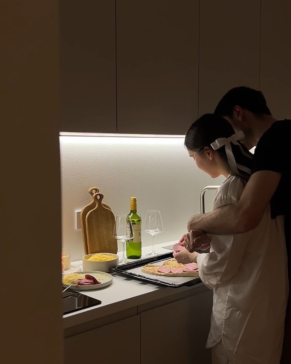 A couple stands close together in a modern kitchen, preparing food on a counter. There are wine glasses, a bottle of wine, pasta, and a cutting board beside them. The lighting is warm and intimate.