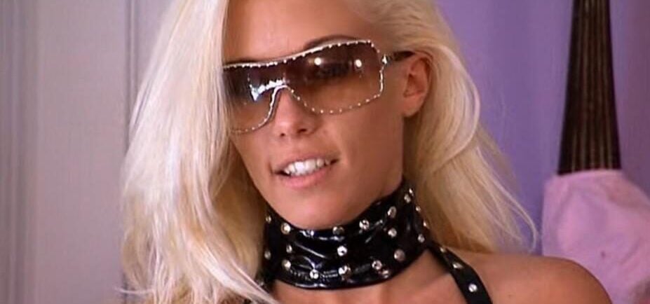 A woman with long platinum blonde hair wears large sunglasses, a studded black choker, and a matching studded top. She is indoors, with a soft purple background behind her.