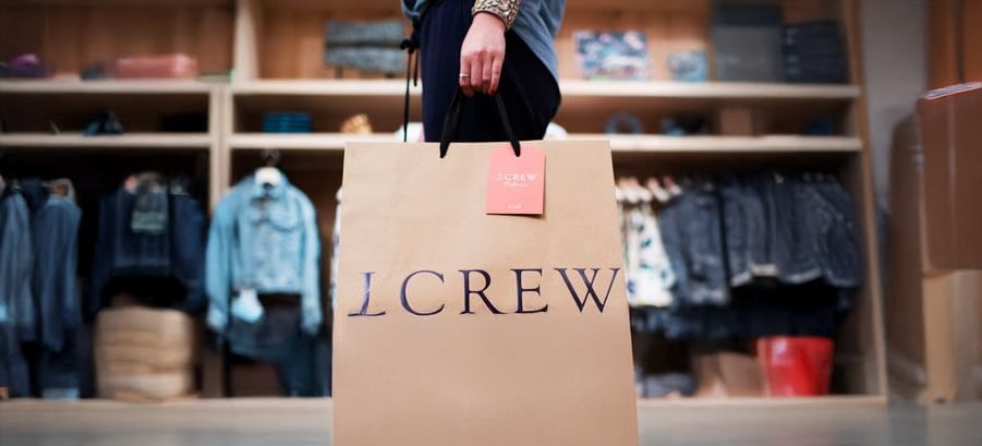 A person holding a large J.Crew shopping bag in a clothing store, with shelves and racks of denim and other apparel visible in the background.