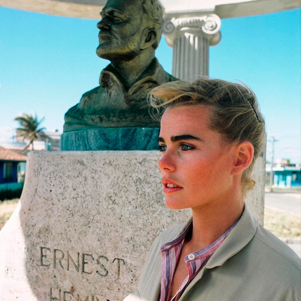 A young woman with blond hair stands in front of a stone monument featuring a bronze bust and the engraved name "Ernest Hemingway," with a bright blue sky and palm trees in the background.