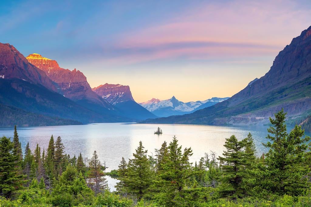 A serene mountain lake surrounded by pine trees, with snow-capped peaks in the background under a pastel sunrise sky. The sun casts a warm glow on the mountain tops.