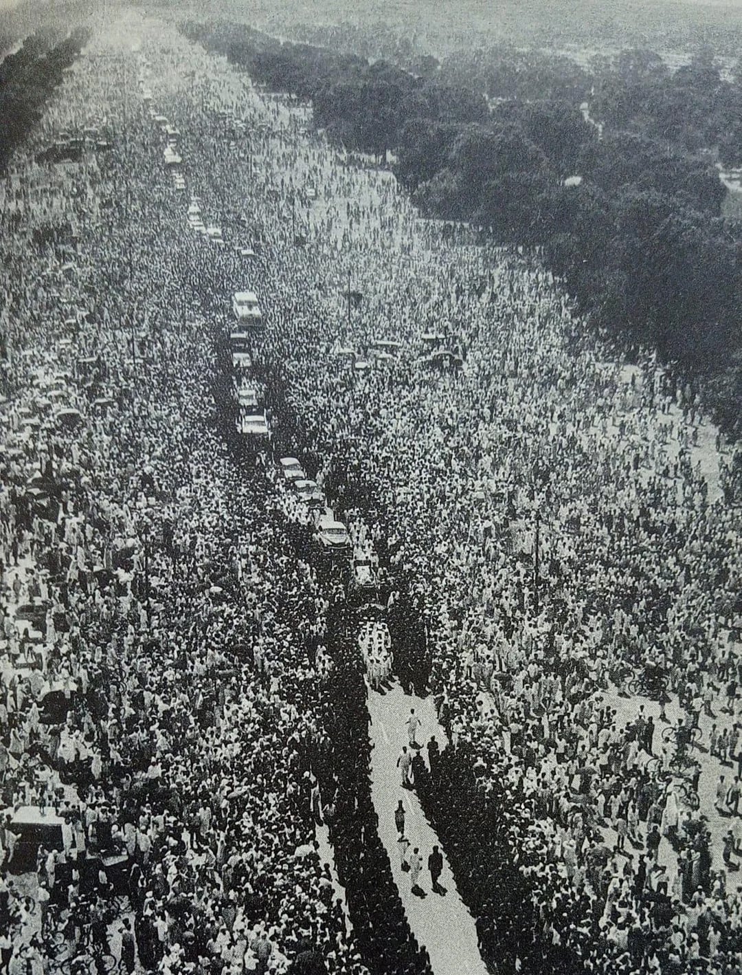 A black and white aerial photo of a dense crowd lining both sides of a wide road for a procession with vehicles and people walking down the center, surrounded by trees in the background.