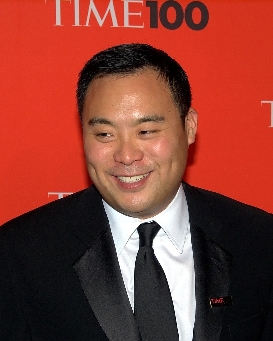 A man in a black tuxedo and tie smiles in front of a red backdrop with "TIME 100" written on it.