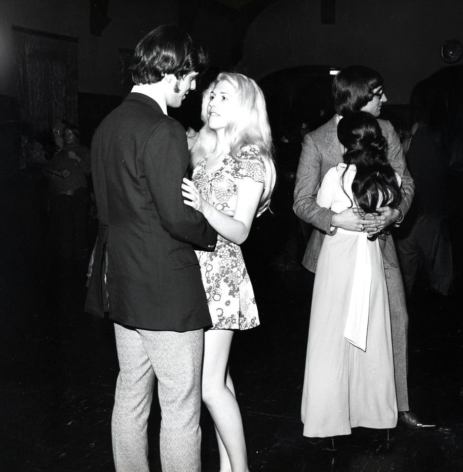 Two couples slow dance in a dimly lit room. The couple in the foreground looks at each other, while the couple in the background hugs closely. The photo is black and white and appears to be from a past decade.