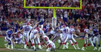 A football game scene showing a kicker from the Buffalo Bills attempting a field goal as New York Giants players jump to block the kick, with the crowd watching in the background.