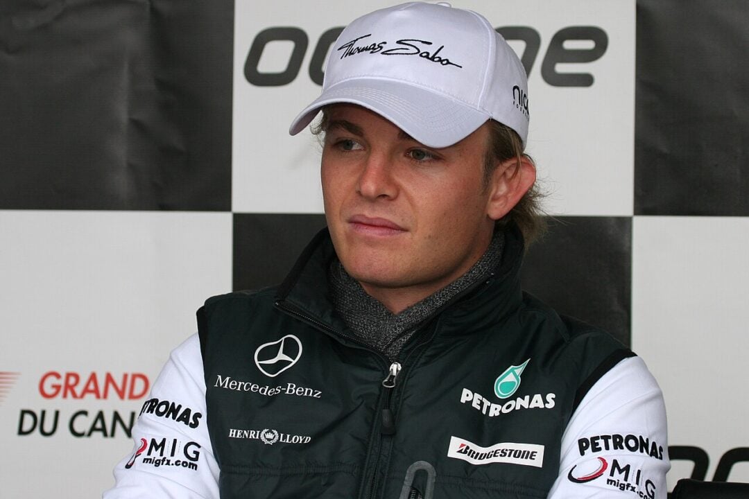 A person in racing team attire and a white cap sits in front of a black-and-white checkered backdrop, likely at a motorsport event or press conference.