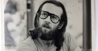 A black-and-white photo of a man with long hair, a full beard, and large glasses. He is wearing a T-shirt and a necklace, looking slightly upwards with an expressive face.