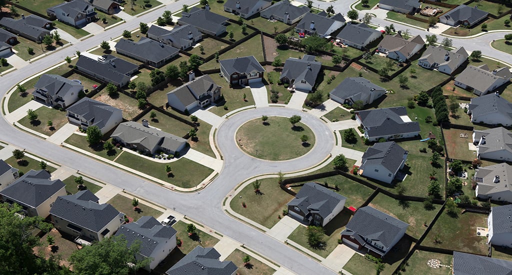Aerial view of a suburban neighborhood featuring a circular cul-de-sac surrounded by single-family homes, lawns, driveways, and tree-lined streets on a sunny day.