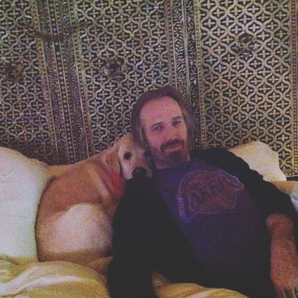 A man with light hair and a beard relaxes on a bed, leaning against a cream-colored dog. They are both resting against white pillows, with an ornate patterned wall in the background.