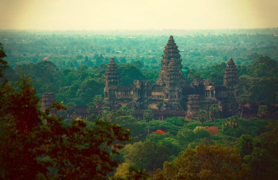A distant view of Angkor Wat temple in Cambodia, surrounded by dense green trees and foliage, with a hazy sky and forested landscape stretching into the background.