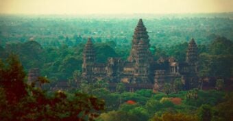 Ancient stone temple complex with tall towers surrounded by lush green trees, set against a hazy landscape in the background.