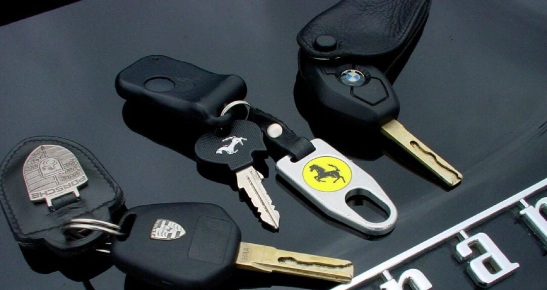 Three car key sets with branded key fobs—Porsche, Ford Mustang, and Ferrari—are placed on the hood of a Ferrari, with the Ferrari emblem and partial logo visible underneath.