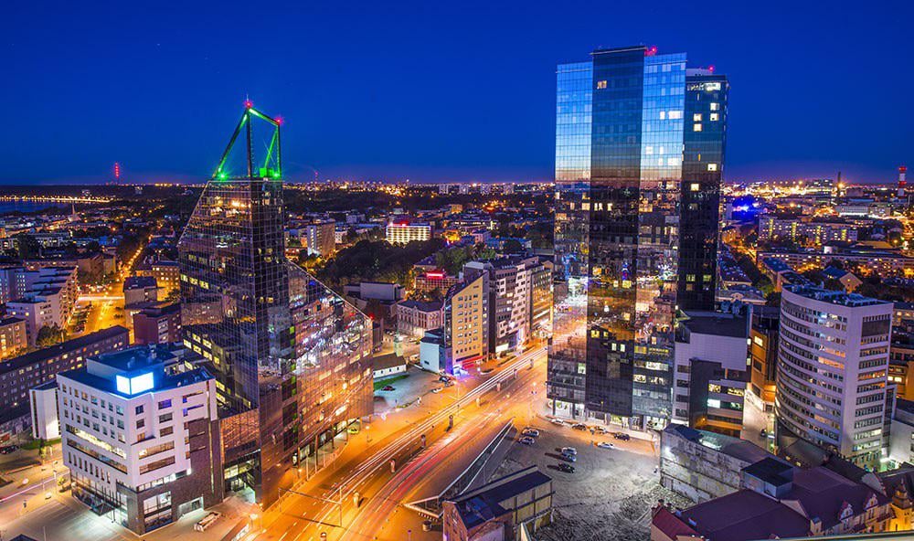 A vibrant cityscape at night with tall modern skyscrapers, brightly lit streets, and buildings against a deep blue sky. City lights create colorful reflections on the glass surfaces of the high-rises.