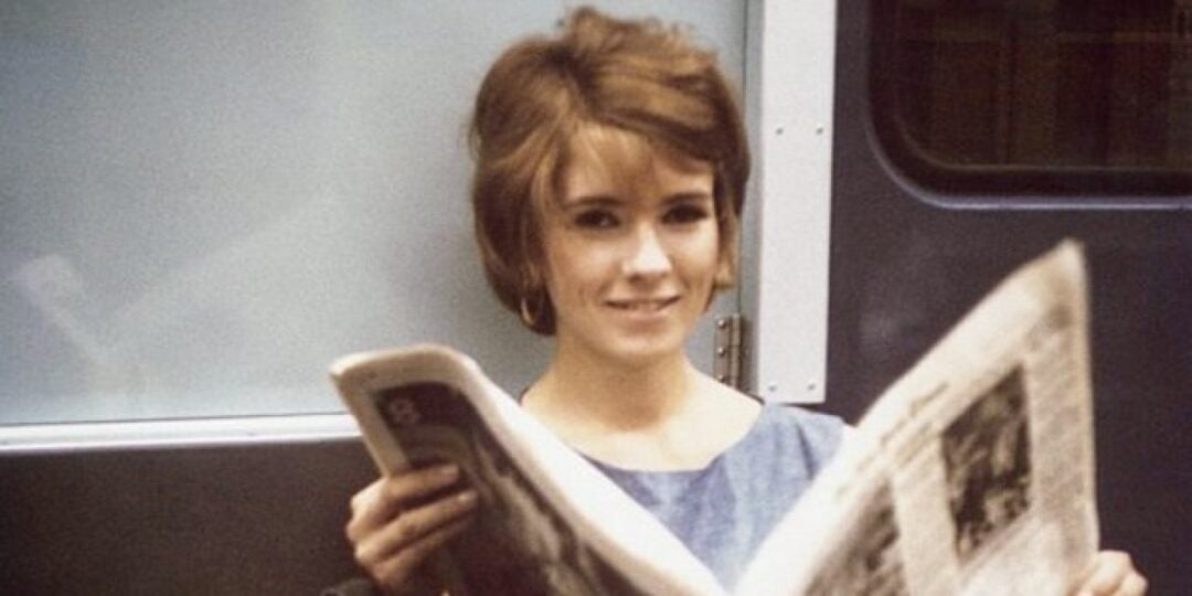 A young woman with short brown hair, wearing a blue top, sits by a window and smiles while reading a newspaper.