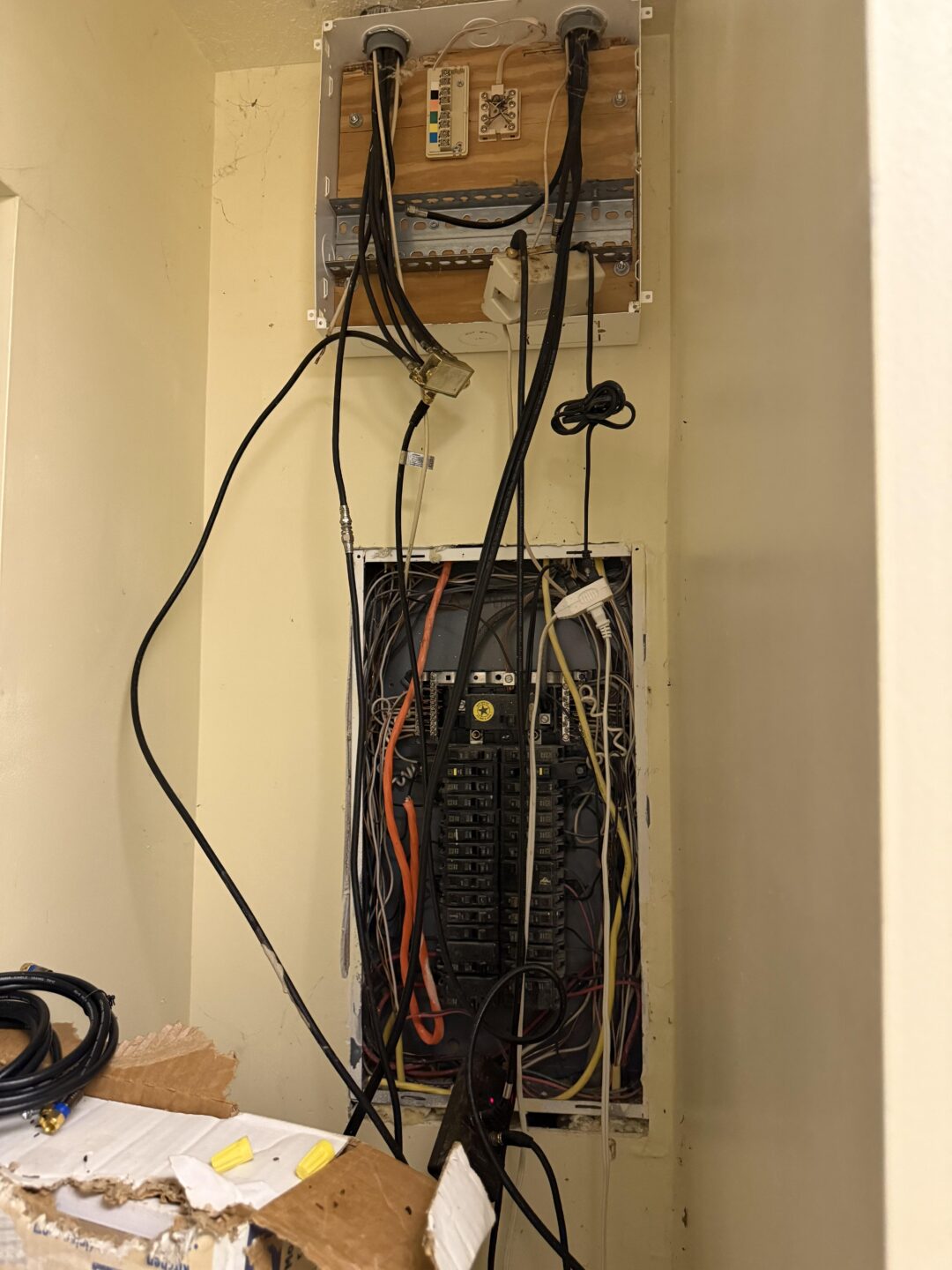 An electrical panel with multiple colored wires and cables hanging loosely, some plugged into outlets and a wooden board above the panel holding more wires. A cardboard box sits at the bottom left of the image.