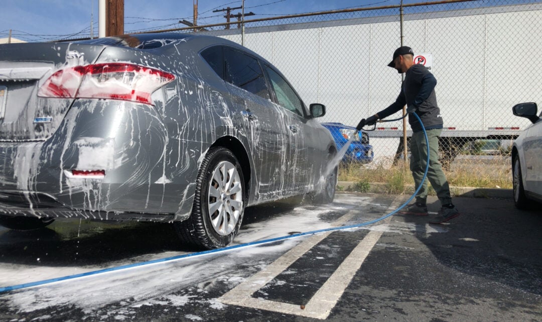 A person uses a hose to rinse soap suds off a silver sedan parked in a lot, with foam covering the car and water spraying. A fence and another car are visible in the background.