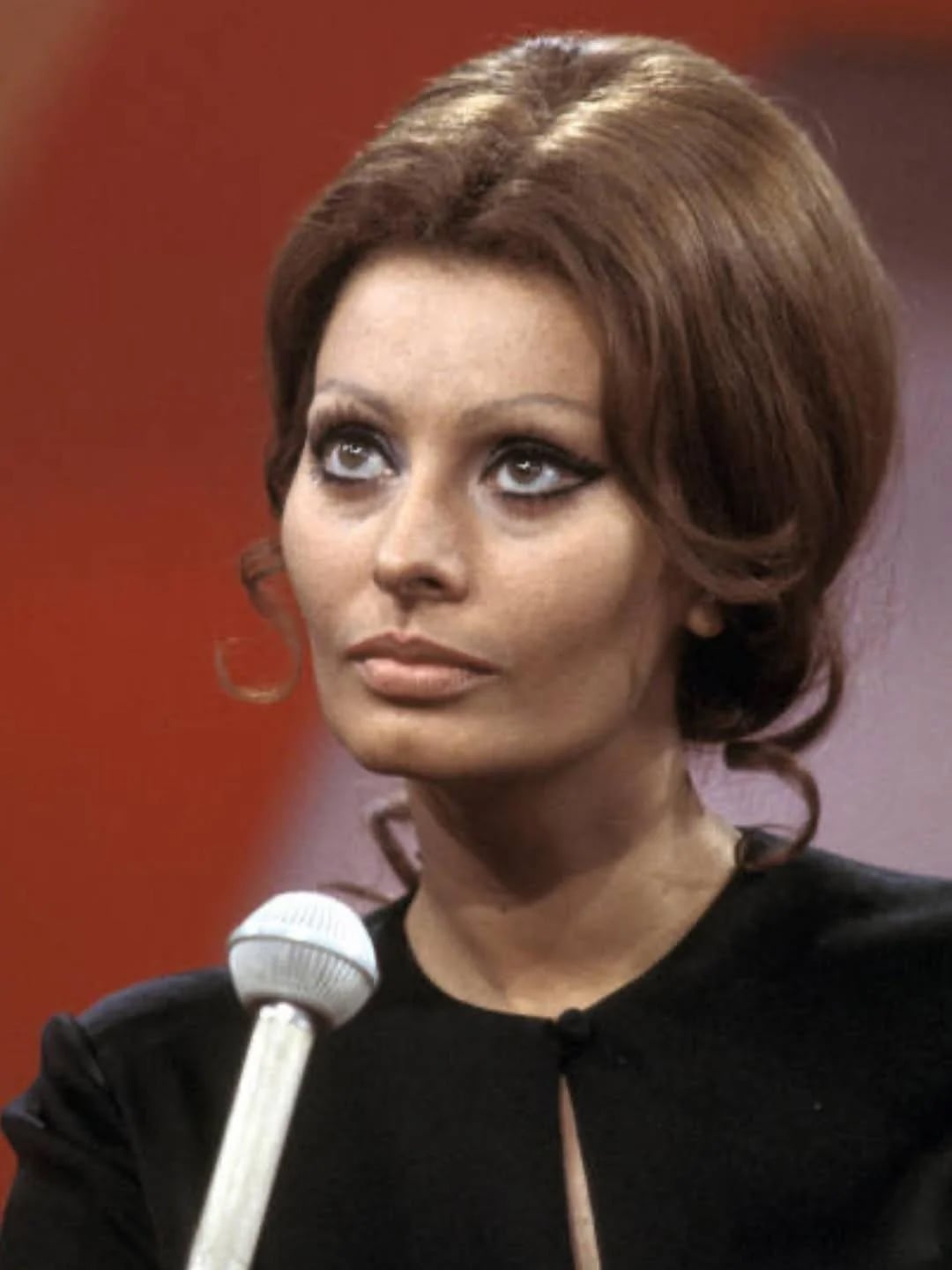 A woman with voluminous brown hair and dramatic eyeliner gazes thoughtfully while seated in front of a microphone, against a blurred warm-toned background.