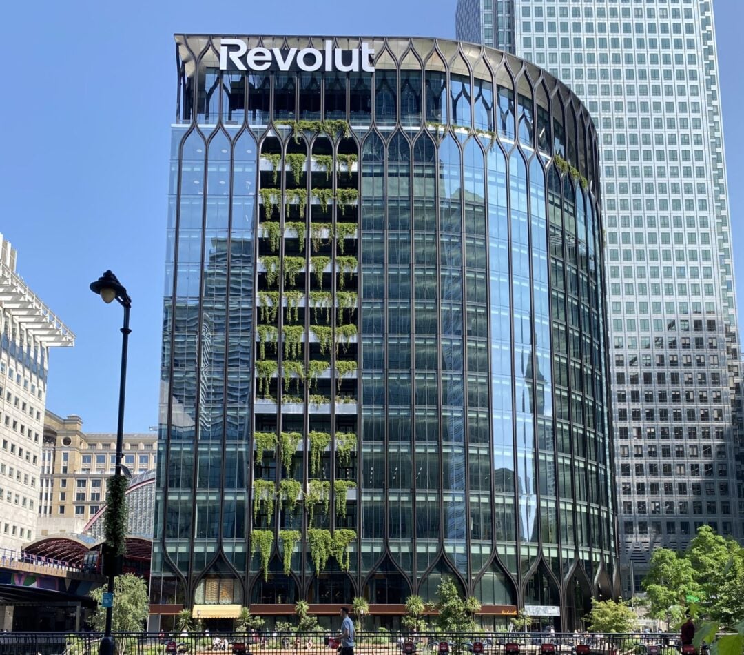 A modern glass office building with the "Revolut" logo on top. Vertical rows of greenery decorate the facade, reflecting nearby skyscrapers under a clear blue sky. People walk and sit in a plaza in front.