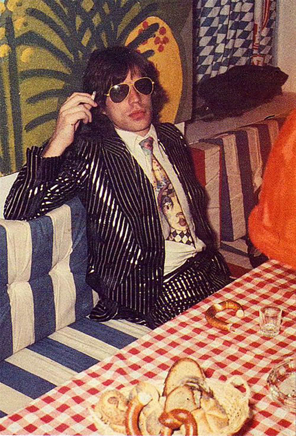 A man in a shiny black pinstripe suit, art-printed tie, and sunglasses sits at a table with a red checkered tablecloth, surrounded by bread and pretzels, holding a cigarette. Bold, colorful fabrics hang in the background.