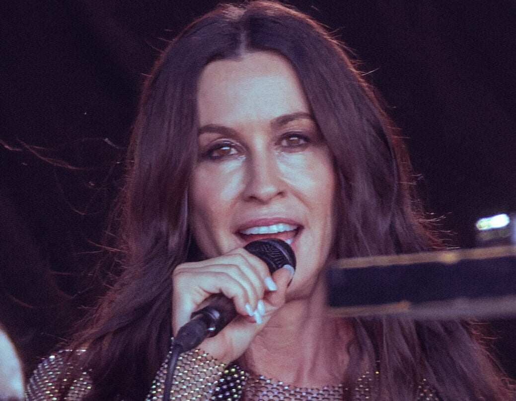 A woman with long brown hair sings into a microphone on stage, wearing a sparkly, sheer top over a black garment, with a dark background behind her.