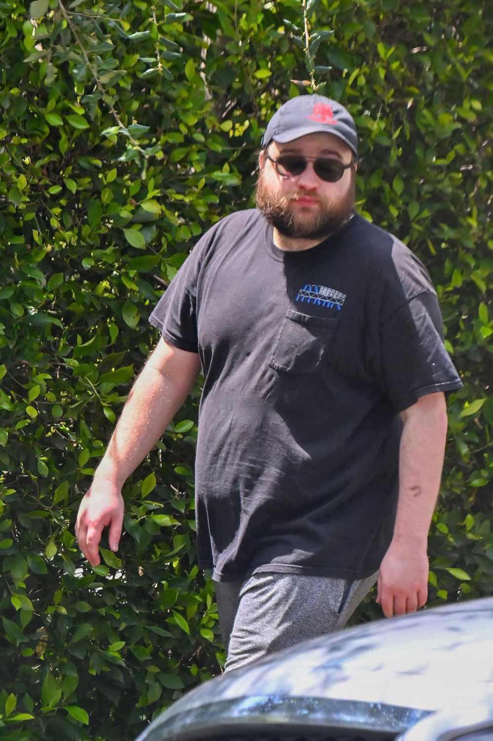 A bearded man wearing a black T-shirt, gray shorts, black cap, and sunglasses walks outdoors in front of dense green bushes on a sunny day.