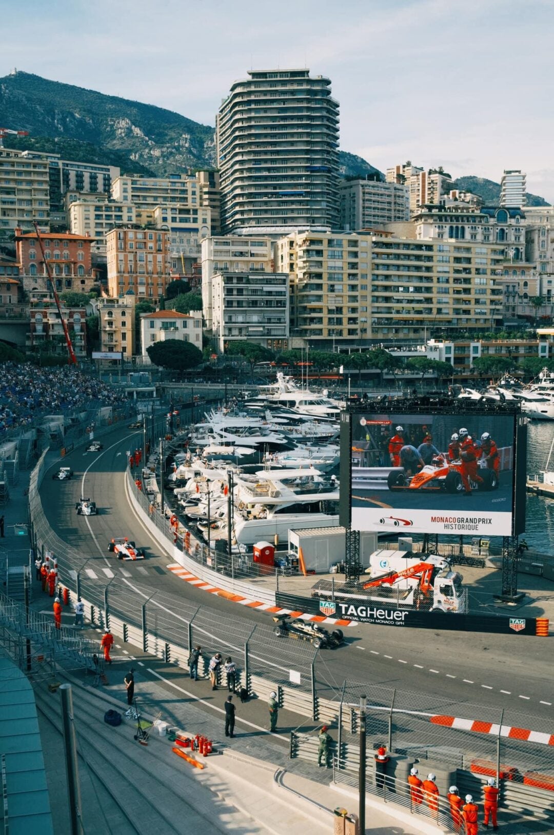 Formula 1 cars race around a tight corner at the Monaco Grand Prix, with yachts docked in the harbor and tall buildings set against a mountain backdrop. Marshals and spectators are visible along the circuit.
