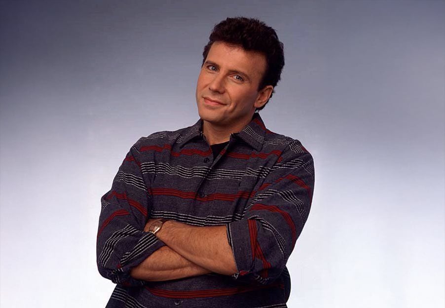 A man with short dark hair, wearing a striped button-up shirt, stands with his arms crossed and smiles slightly against a plain light background.