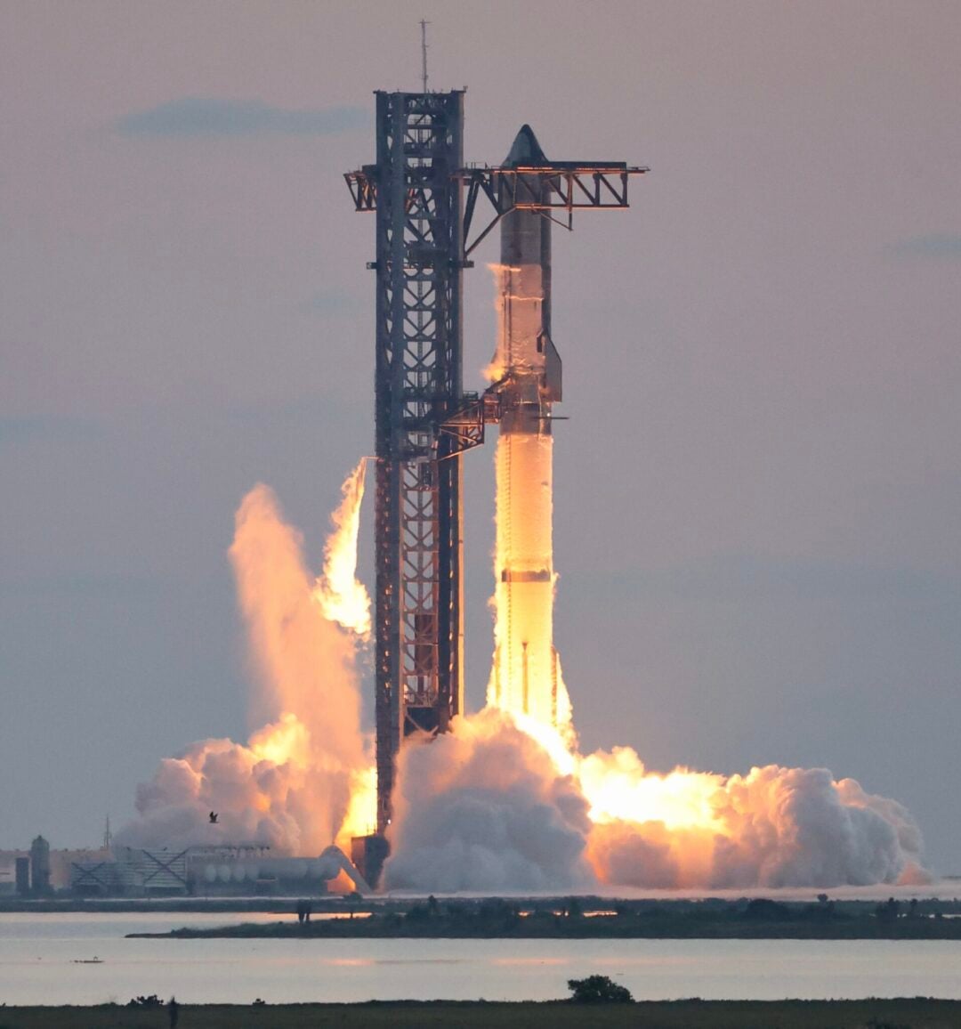 A rocket lifts off from a launch pad, emitting bright flames and smoke at its base, with a metal tower structure beside it. The sky is mostly clear, and the scene is reflected in the water below.