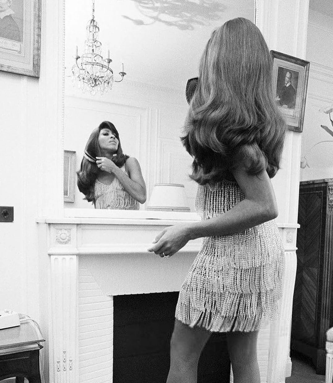A woman in a fringed dress stands at a fireplace, looking at her reflection while fixing her hair in a large mirror in an elegant room with a chandelier and framed portraits.