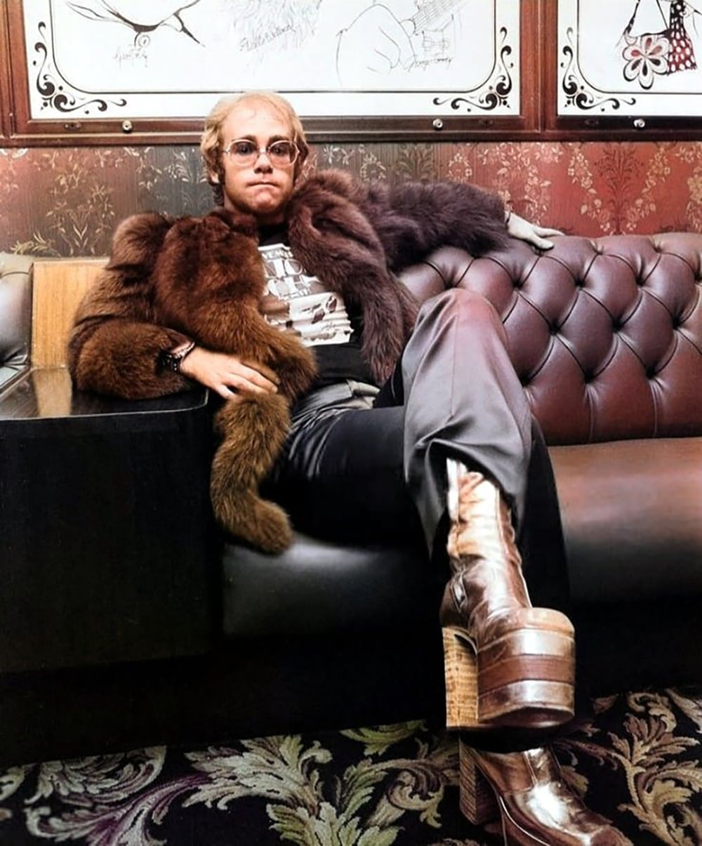 A person in sunglasses and a brown fur coat lounges on a tufted leather sofa, wearing shiny pants and tall platform boots. The background features patterned wallpaper and framed drawings.