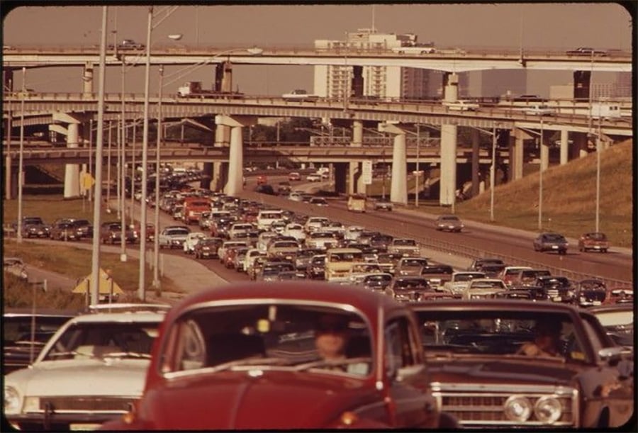 Heavy traffic on a multi-lane freeway with many cars backed up. Several overpasses and elevated roads intersect above, with tall buildings visible in the background under a hazy sky.