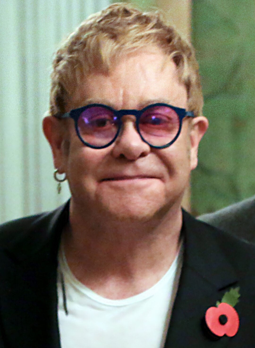 A person with short, light brown hair, wearing blue-rimmed glasses, a hoop earring, a white shirt, and a dark blazer with a red poppy pin, smiles at the camera.