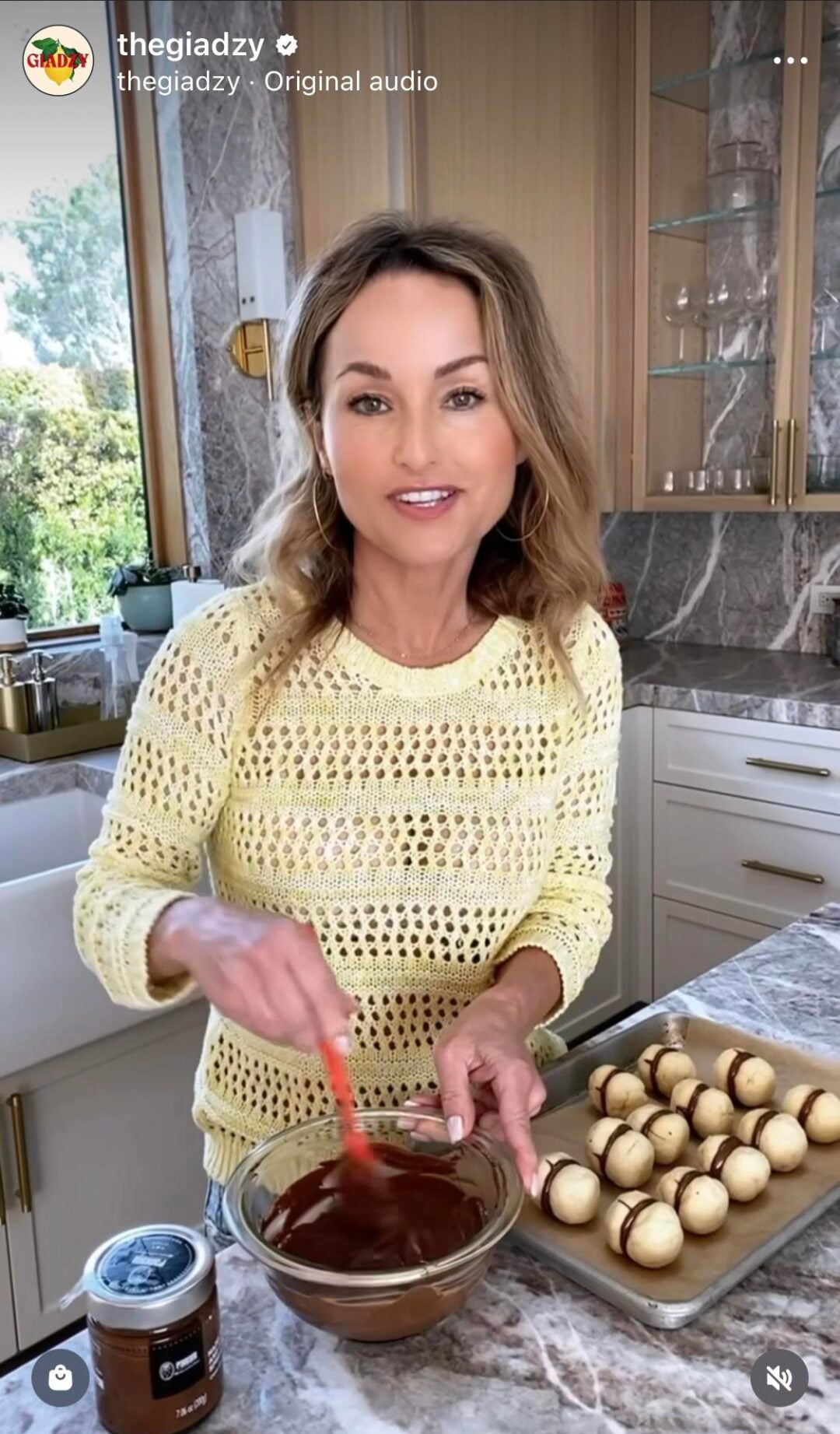 A woman in a yellow sweater stirs melted chocolate in a glass bowl in a modern kitchen. On the counter are a jar of chocolate spread and a tray of round sandwich cookies.