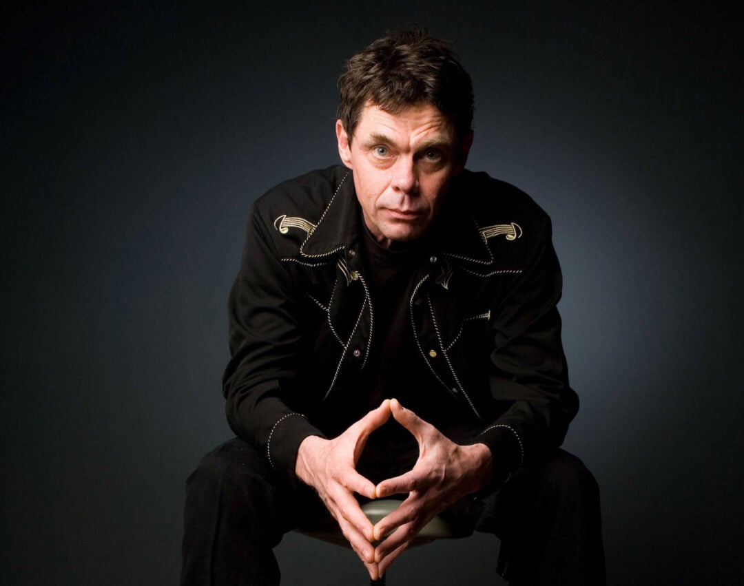 A man with short dark hair sits on a stool, leaning forward with his elbows on his knees and his hands clasped. He wears a black jacket with decorative stitching over a dark shirt, against a dark background.