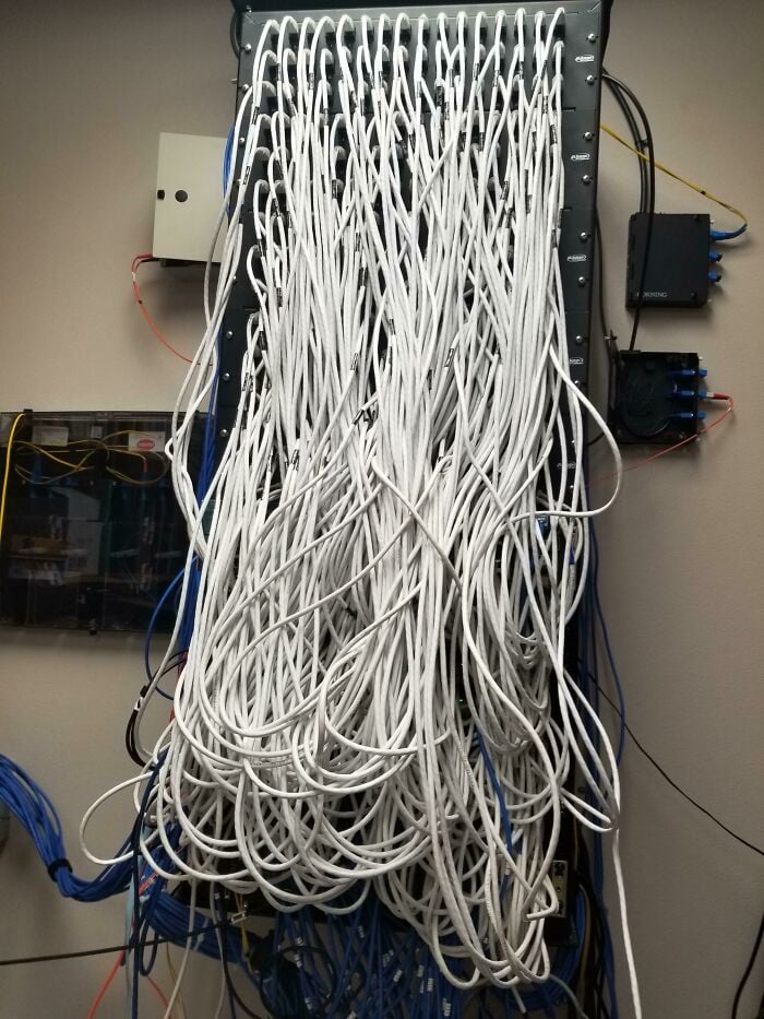 A network rack with a chaotic tangle of white Ethernet cables hanging down, creating a cluttered and disorganized appearance. Some blue cables are also visible at the bottom and sides of the rack.