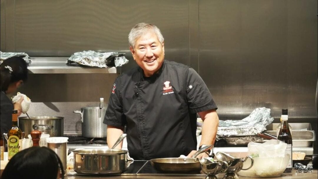 A smiling chef in a black uniform stands in a commercial kitchen, surrounded by cooking pots, bottles, and utensils, with prepared food and ingredients on the counters.