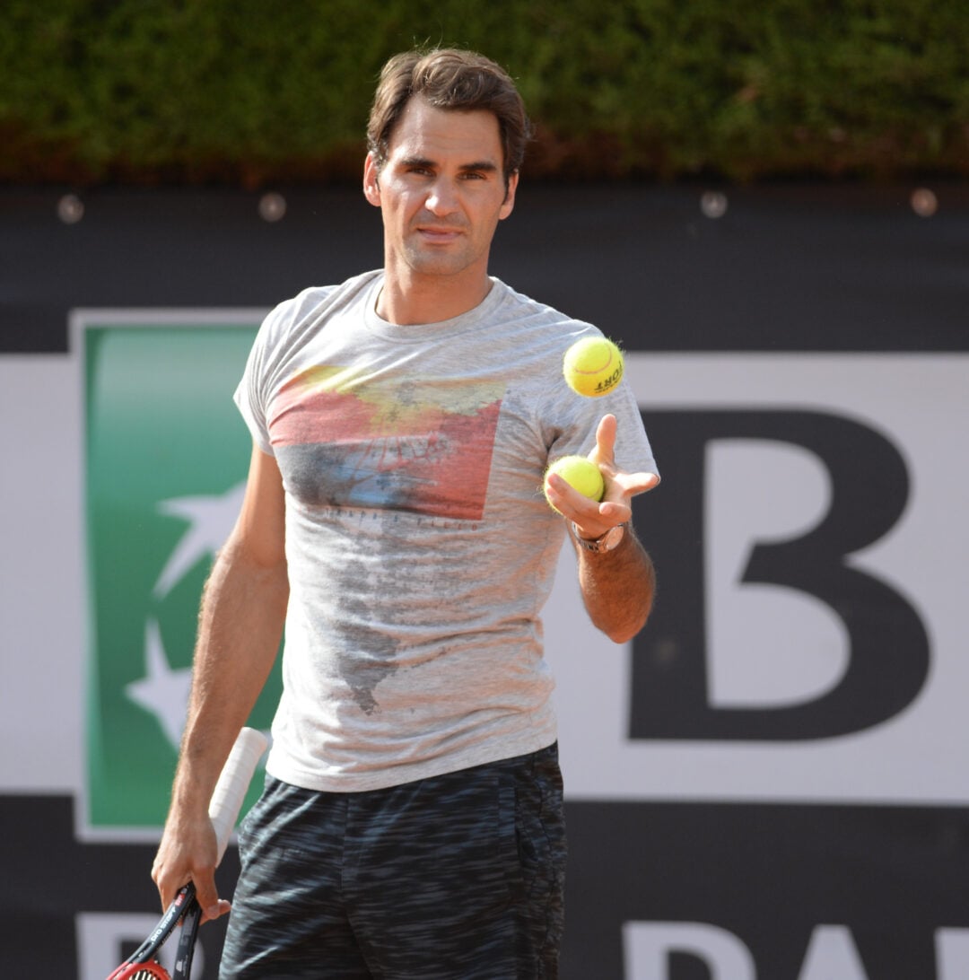 A man in a gray T-shirt and black shorts is tossing a tennis ball with one hand while holding a tennis racket and another ball in the other hand on an outdoor tennis court.
