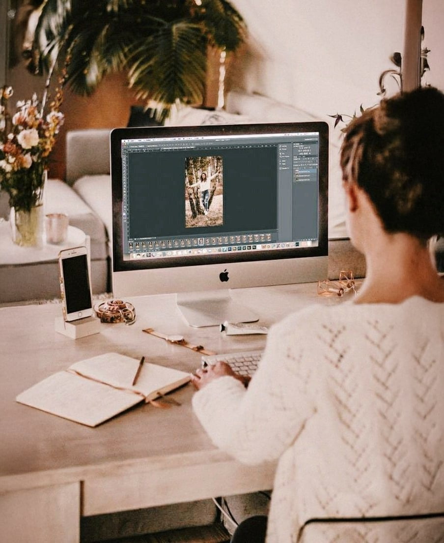 A person sits at a desk using a desktop computer to edit a photo. The workspace includes a notebook, smartphone, and flowers, with warm lighting and plants in the background.