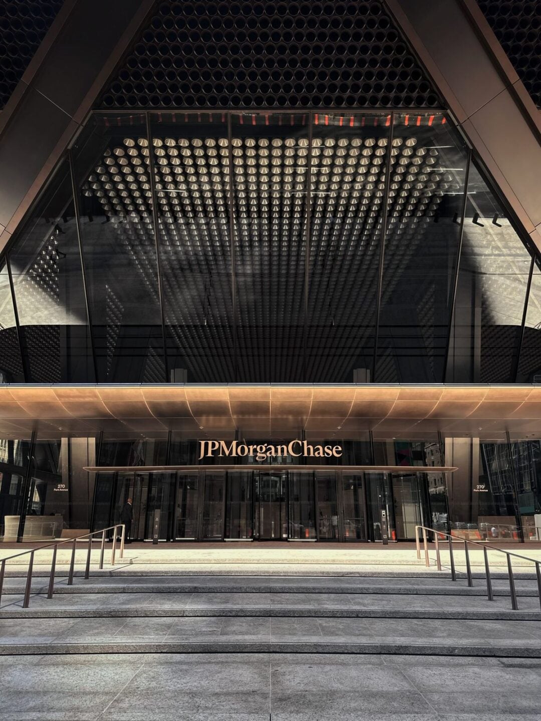 The image shows the main entrance of a modern glass building, with "JPMorgan Chase" written above the doors. Reflections and geometric patterns are visible on the glass facade. Steps and railings lead up to the entryway.